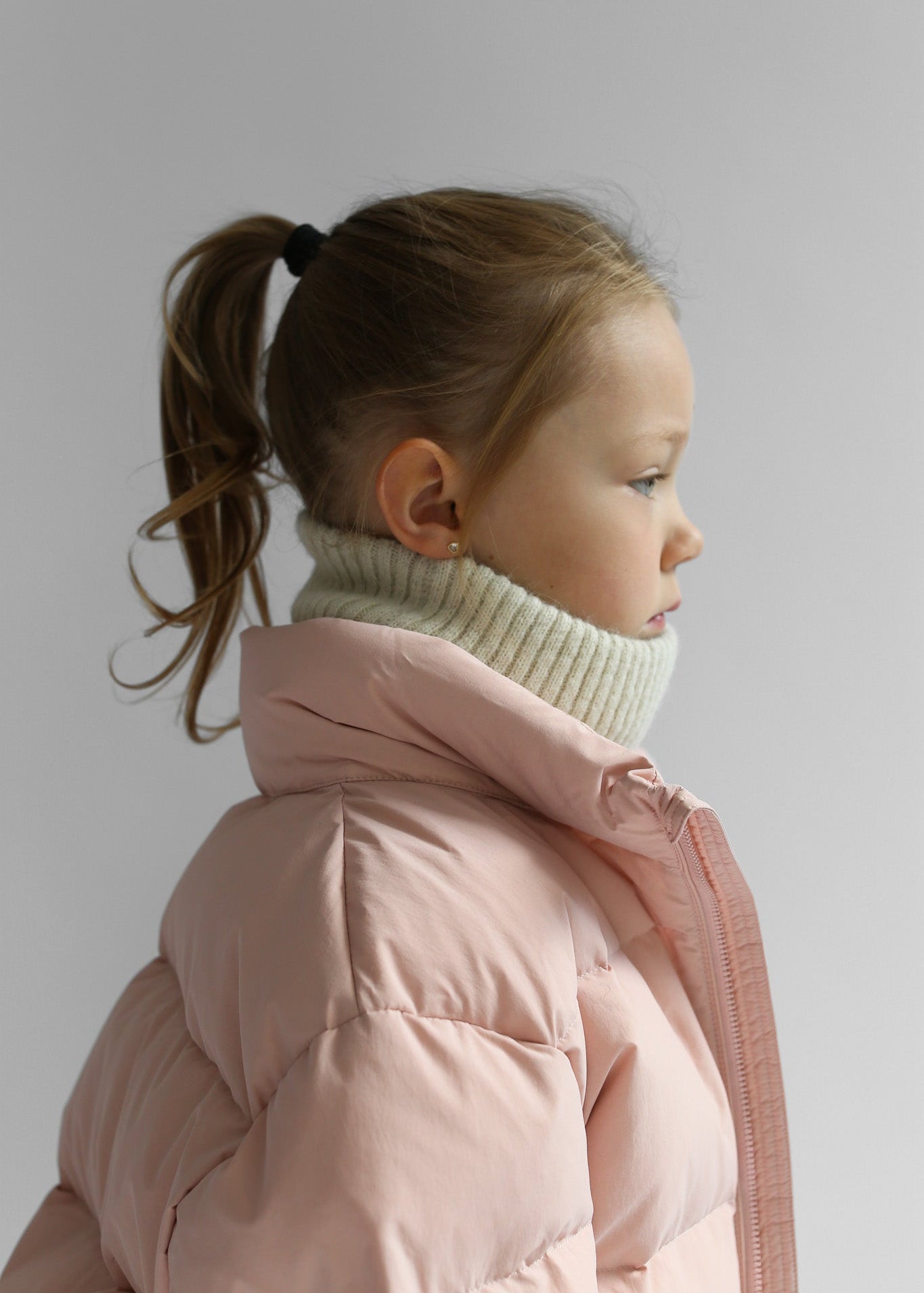 Child wearing a pink puffer jacket and white neck warmer against a plain background