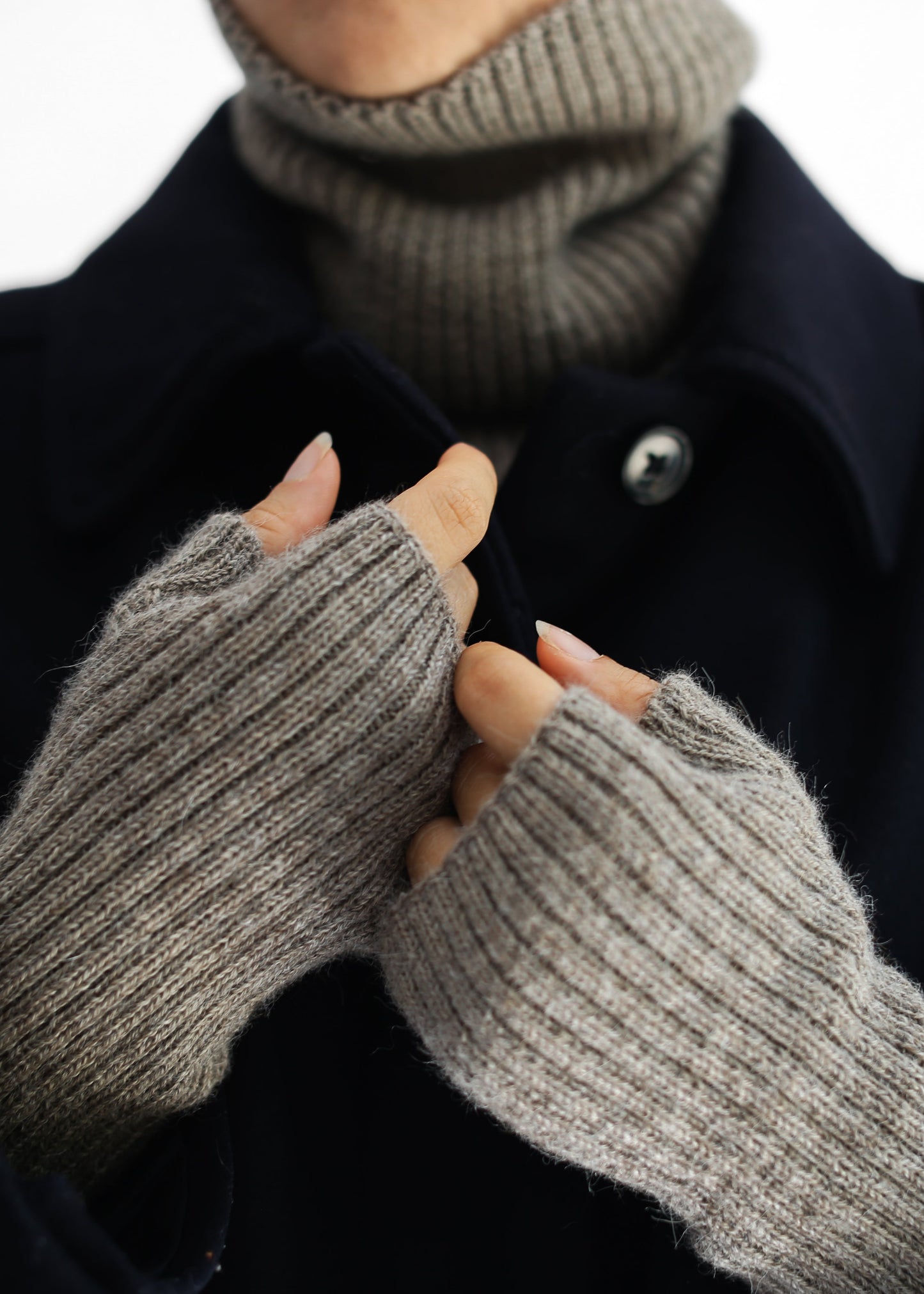 Close-up of a person wearing gray ribbed knit fingerless gloves over a dark navy coat and taupe turtleneck sweater, against a plain white background.
