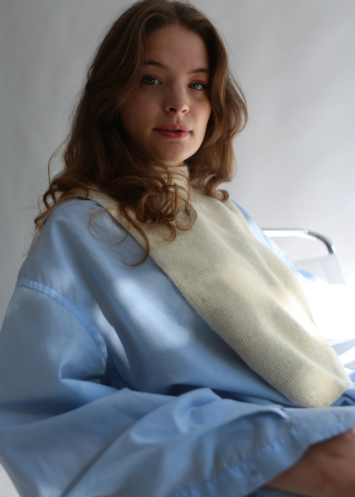 A woman with wavy light brown hair wearing a light blue hospital-style gown with a cream knit sweater draped over her shoulders, photographed against a plain light gray background.