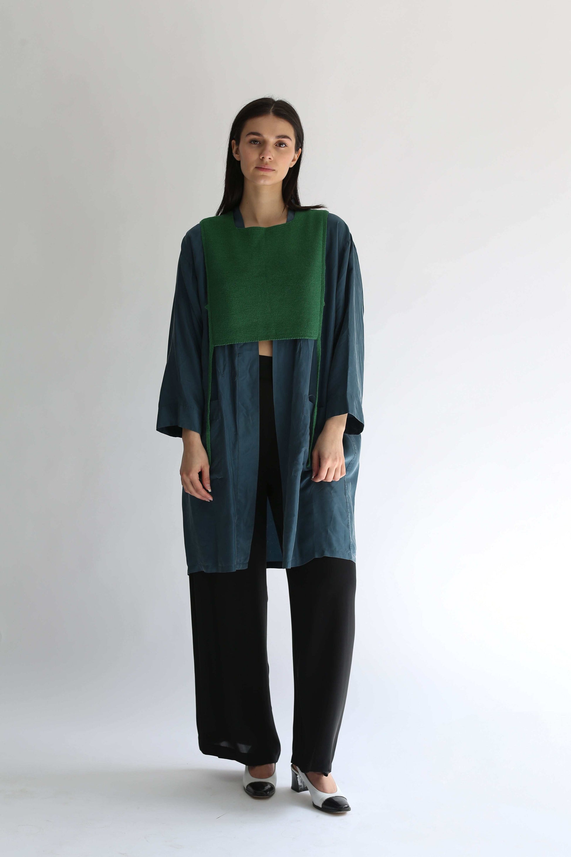 A woman wearing a green knit apron with black pants and long blue shirt on a plain background