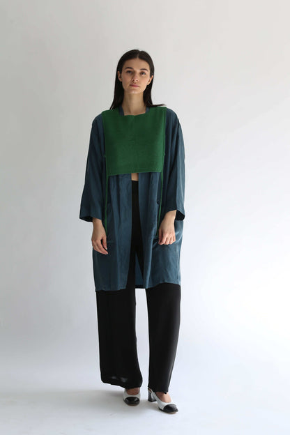 A woman wearing a green knit apron with black pants and long blue shirt on a plain background