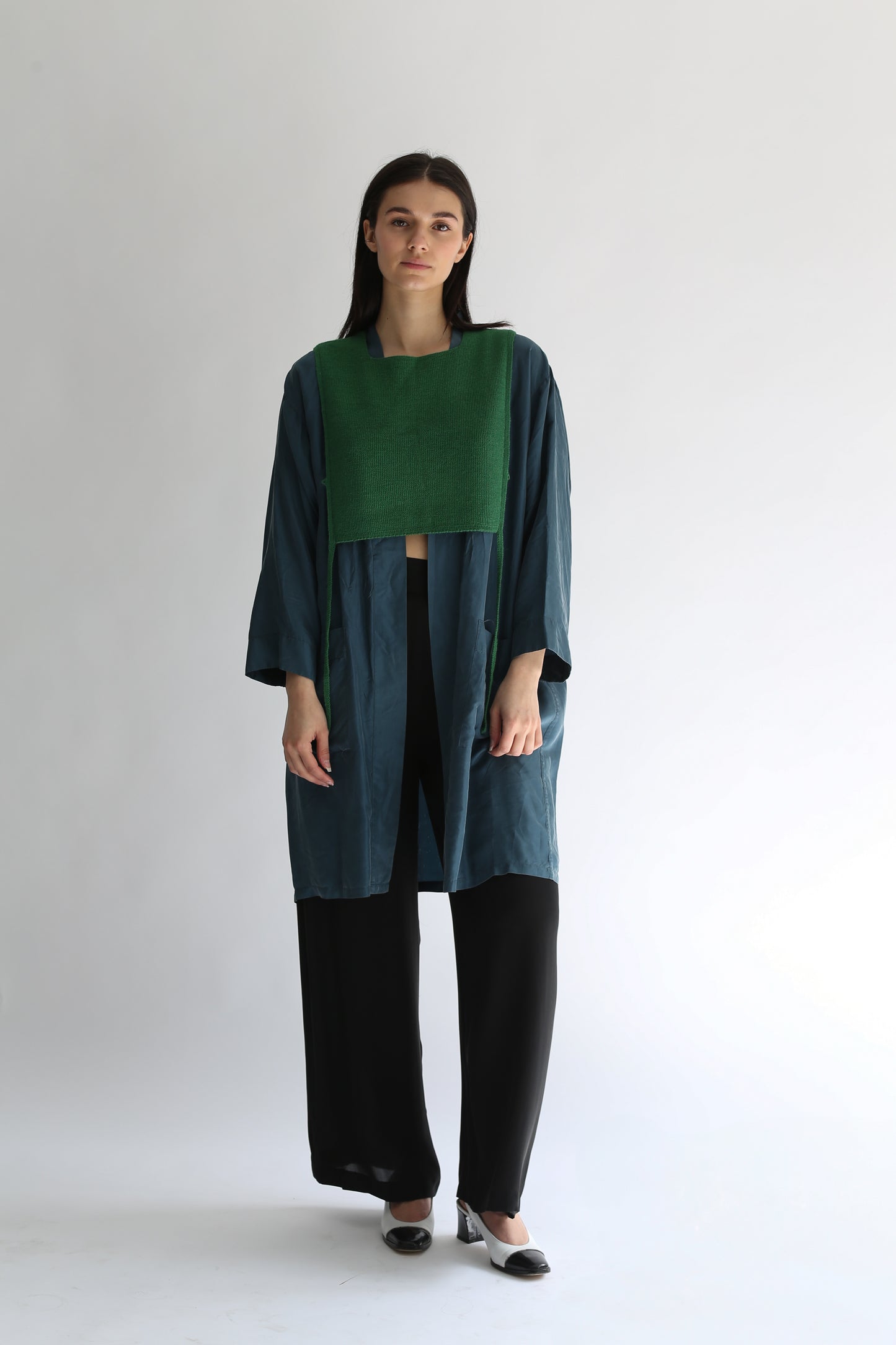 A woman wearing a green knit apron with black pants and long blue shirt on a plain background