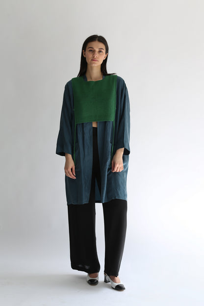 A woman wearing a green knit apron with black pants and long blue shirt on a plain background