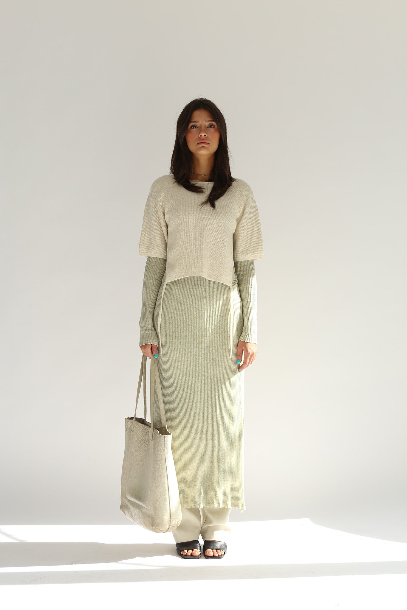 Model wearing the belacaro apron with sleeves over a long ribbed knit dress, carrying a beige leather tote bag.