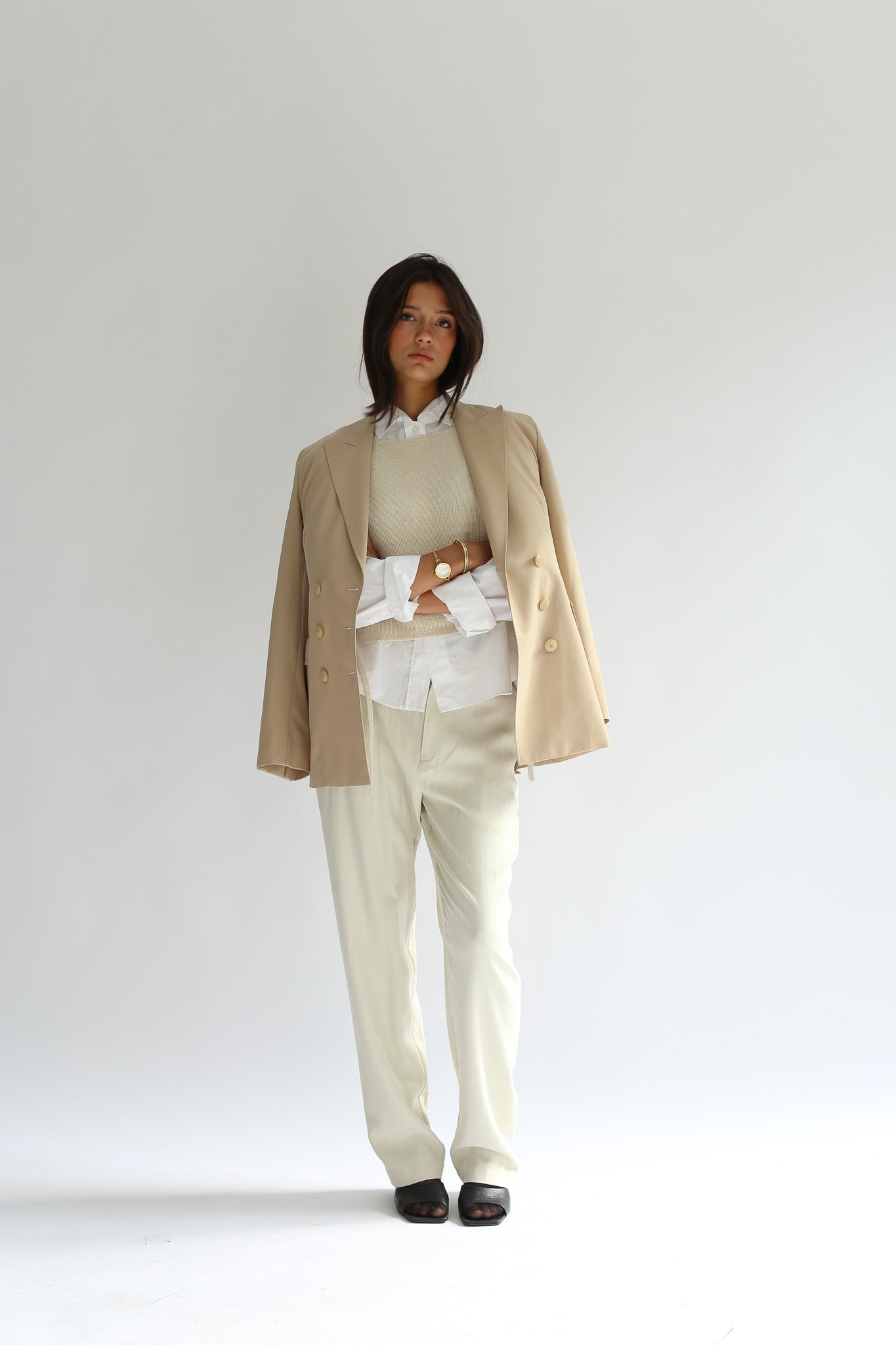 Model wearing the belacaro apron with sleeves knit layered over a white shirt and beige blazer, styled with cream trousers and black sandals, crossed arms