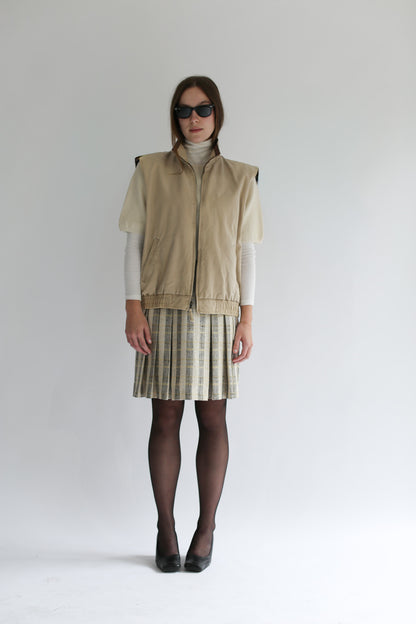 Model wearing the beige belacaro apron with sleeves knit layered over a turtleneck and pleated skirt, paired with black heels and sunglasses.