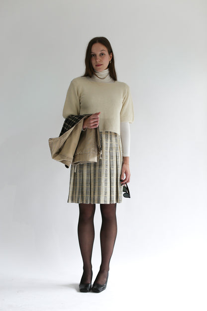 Model holding a beige jacket and sunglasses, wearing the belacaro apron with sleeves knit over a plaid pleated skirt and white turtleneck.