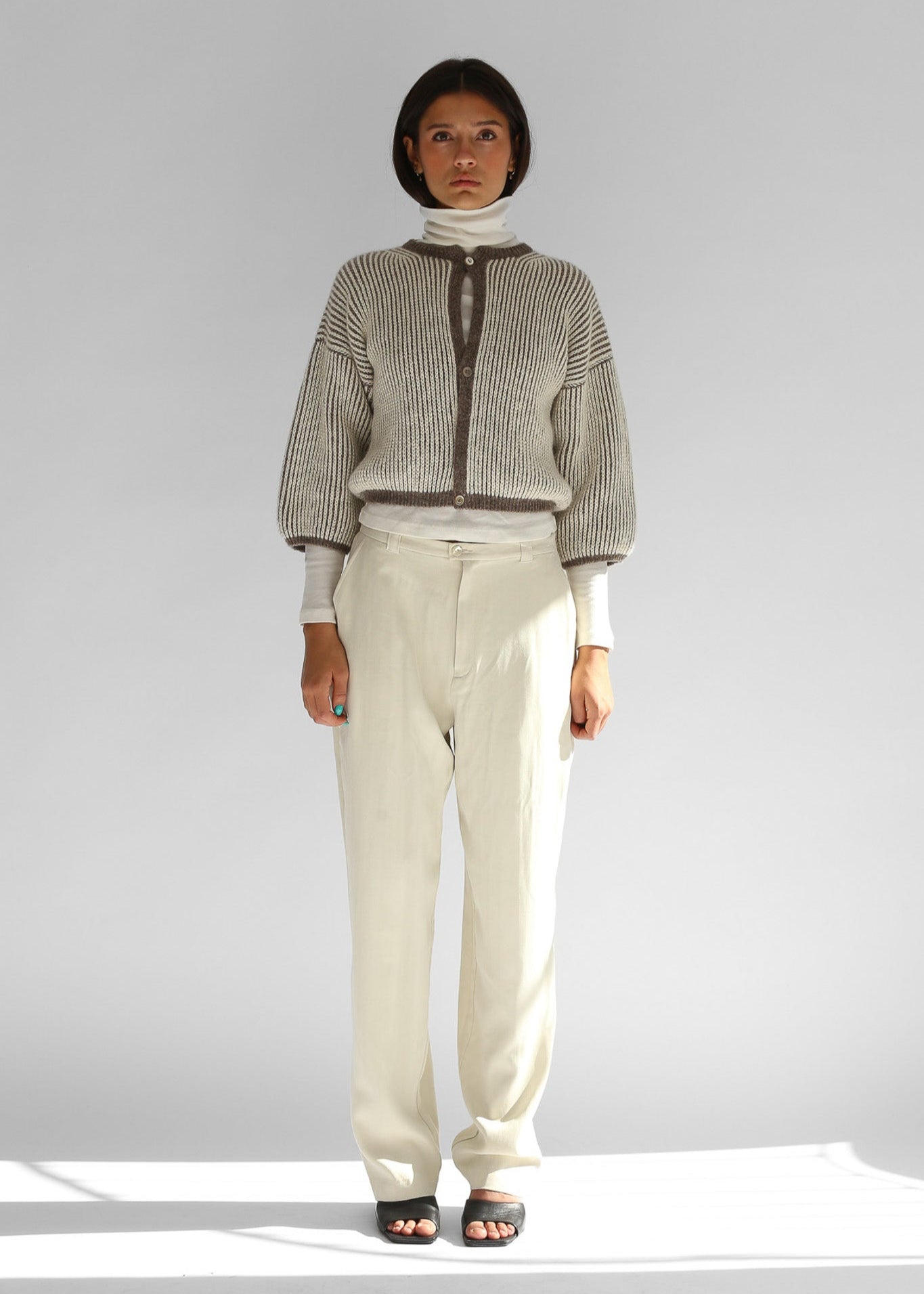 Model wearing a knitted brown and white bicolor cardigan and beige pants on a plain background