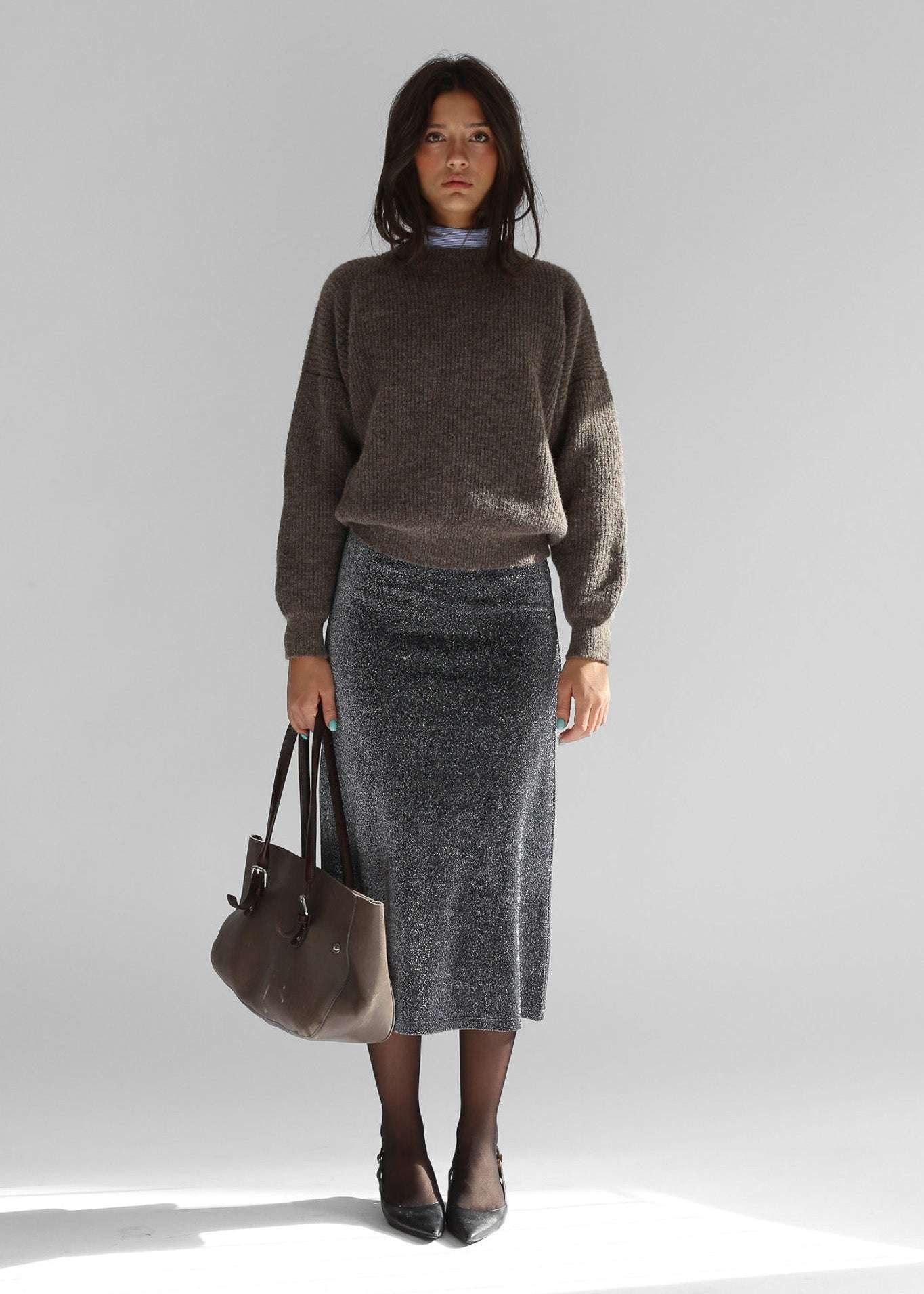 A woman wearing a brown crewneck sweater with a grey skirt on a plain background