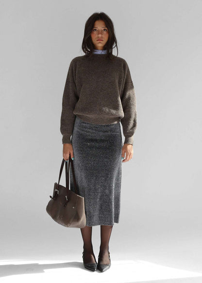A woman wearing a brown crewneck sweater with a grey skirt on a plain background