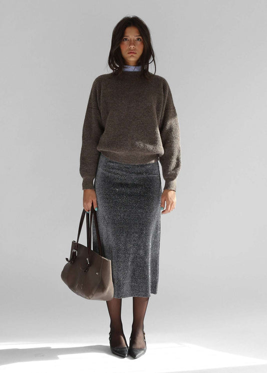 A woman wearing a brown crewneck sweater with a grey skirt on a plain background