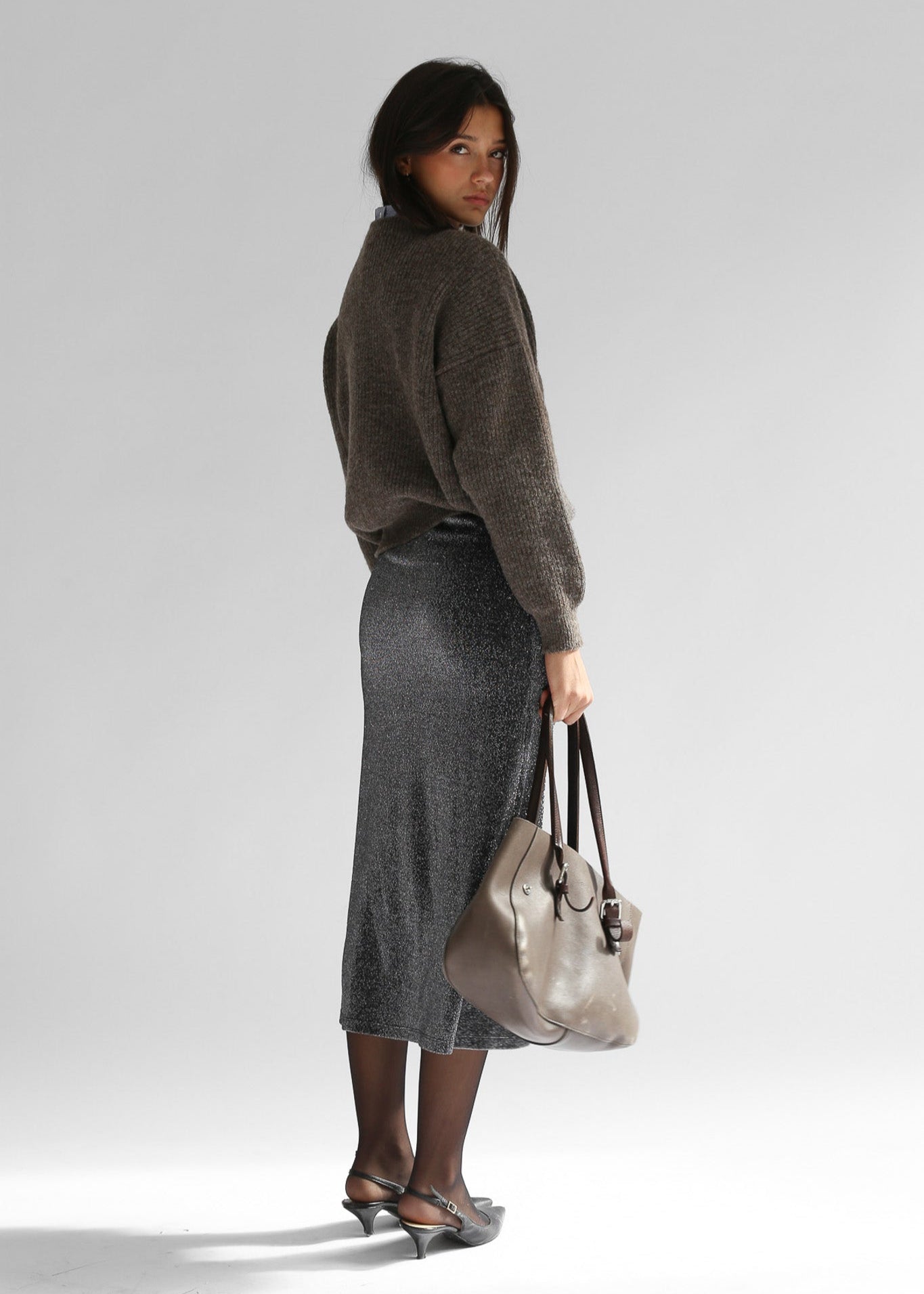 A woman wearing a brown crewneck sweater with a grey skirt on a plain background from the back