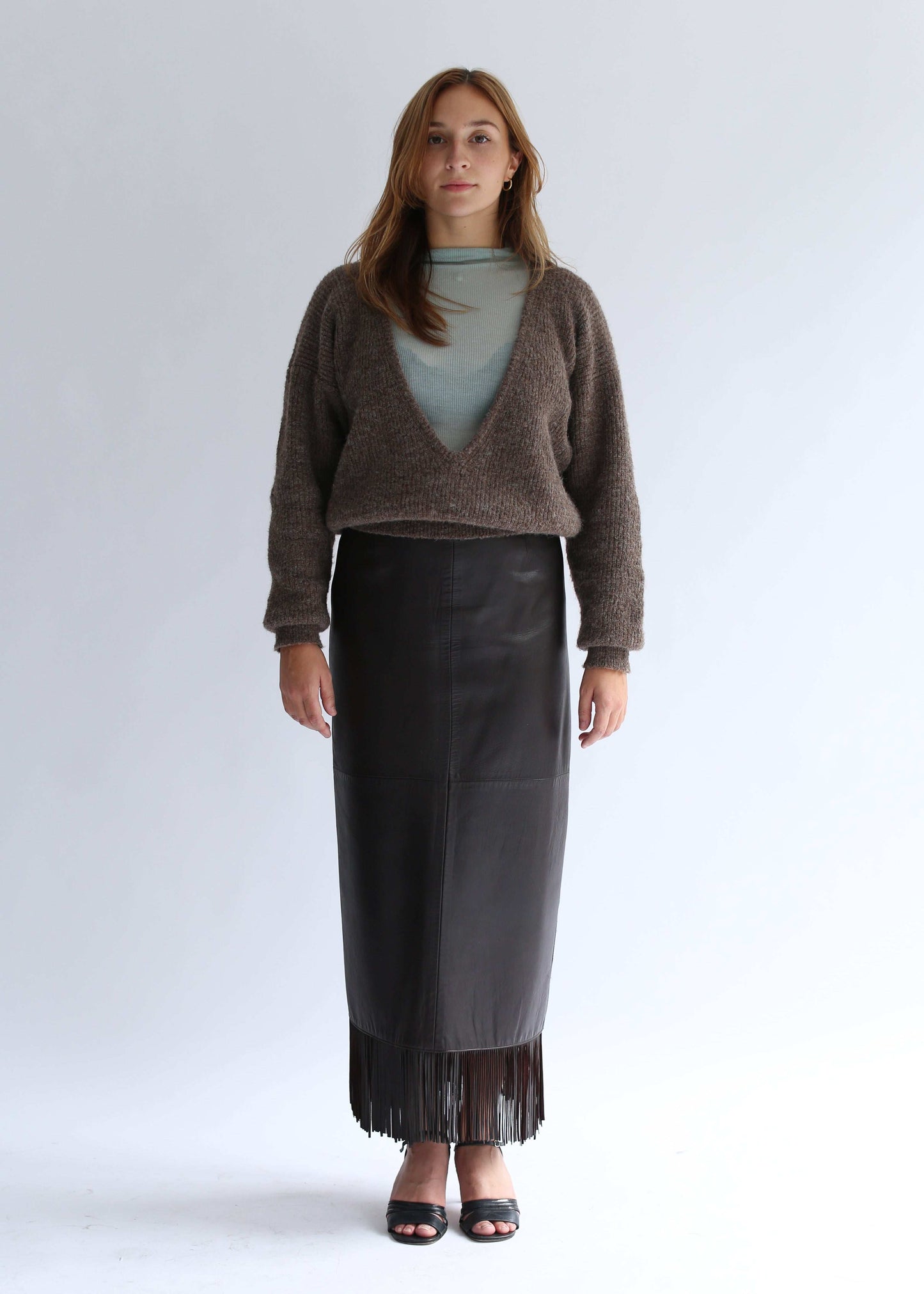 Woman wearing a brown sweater with a deep v neck, light top and a long, fringed lack leather skirt