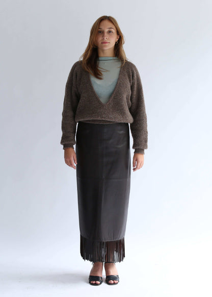 Woman wearing a brown sweater with a deep v neck, light top and a long, fringed lack leather skirt