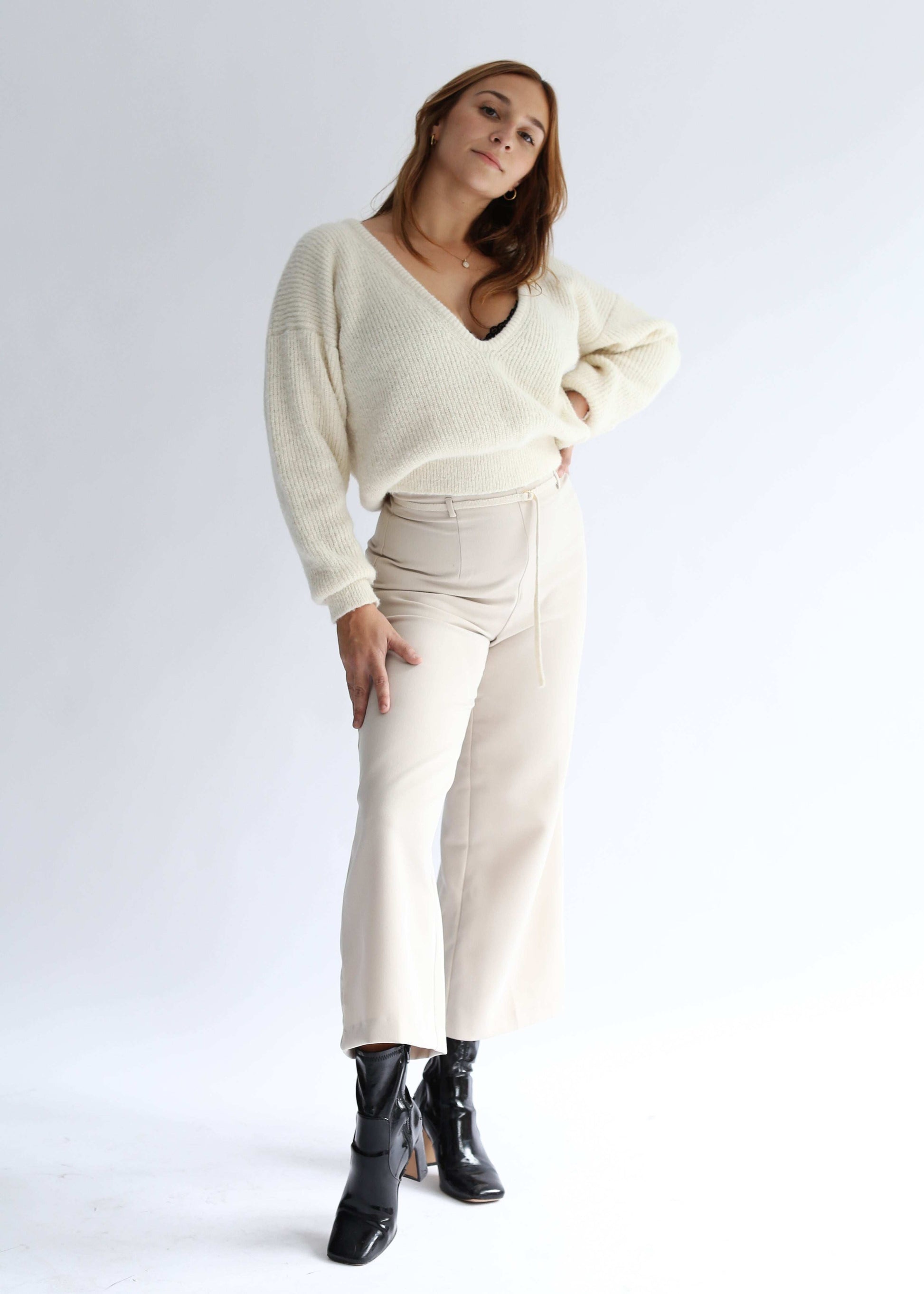 A woman standing, wearing a white sweater with a deep v neck and white pants over a white background