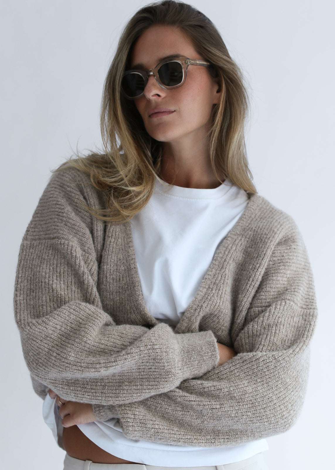 A woman wearing a chunky knit beige cardigan over a white t-shirt and sunglasses, posing with her arms crossed against a plain light gray background.