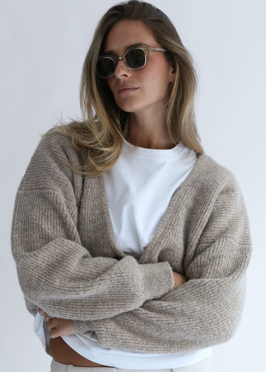 A woman wearing a chunky knit beige cardigan over a white t-shirt and sunglasses, posing with her arms crossed against a plain light gray background.