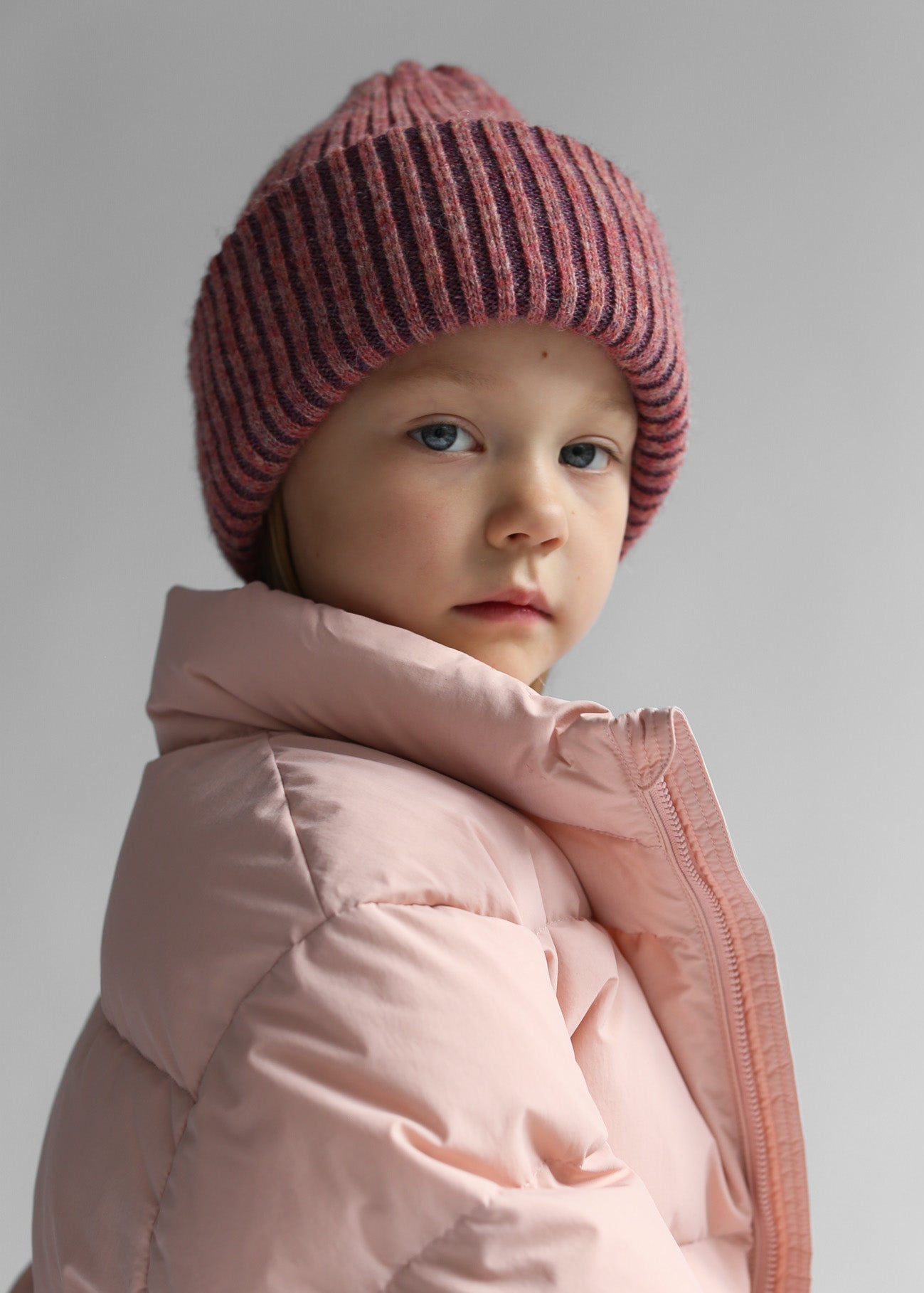 Child wearing the Belacaro kids beanie in heather pink baby alpaca, styled with a soft pink puffer jacket, side portrait view.