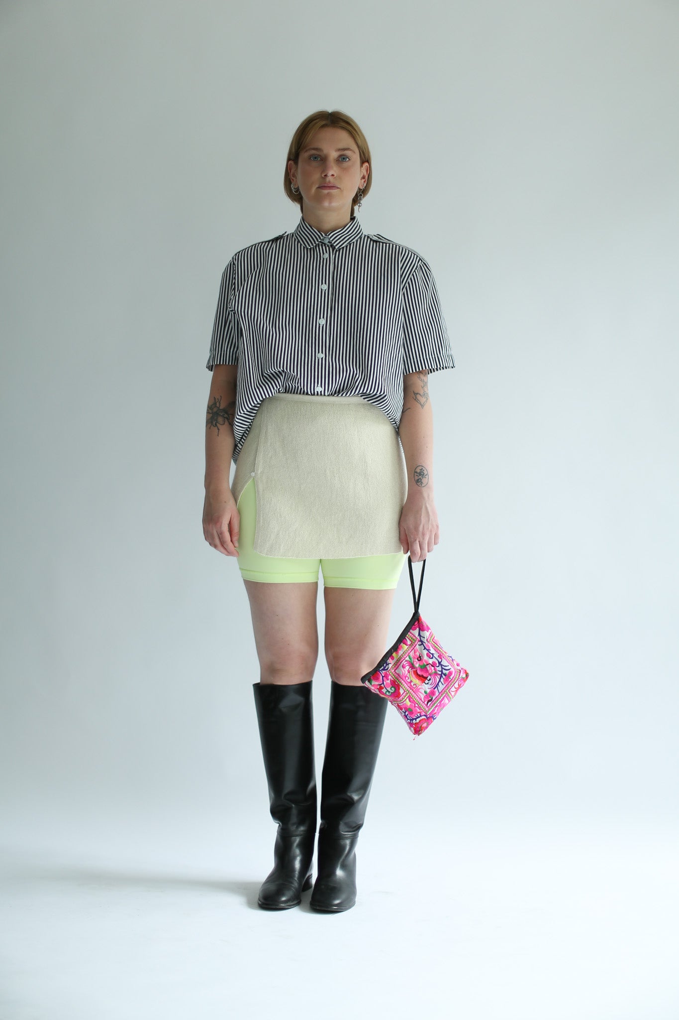 Woman wearing a white knit top over a striped shirt, light green shorts, black knee-high boots, and holding a colorful bag on a white background