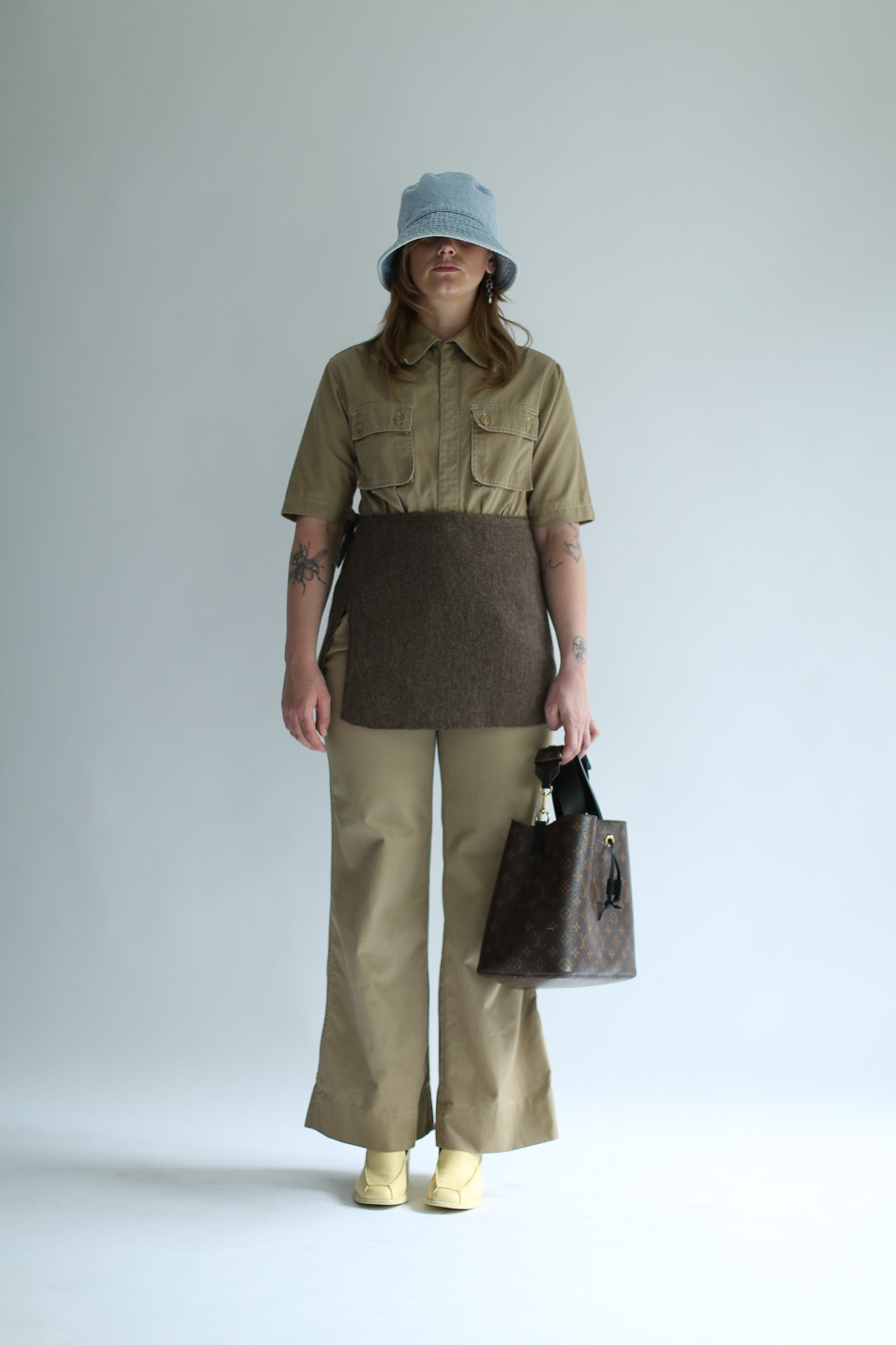 Person wearing a brown knit skirt over beige shirt and beige pants holding a brown bag against a white background