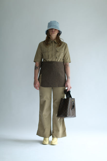 Person wearing a brown knit skirt over beige shirt and beige pants holding a brown bag against a white background