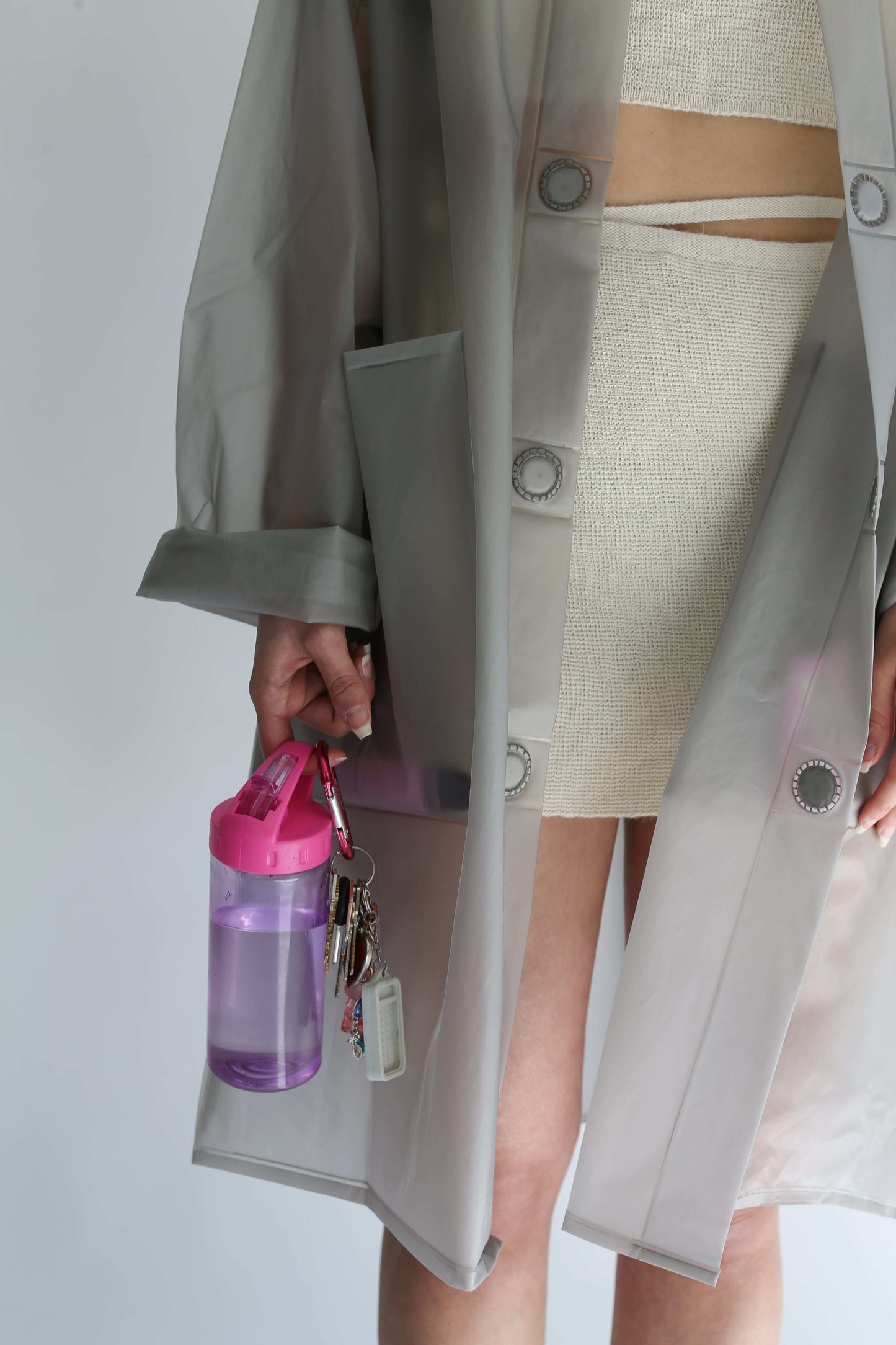 Detail of a woman wearing a transparent trench coat over a white knit skirt, holding keys and a bottle 