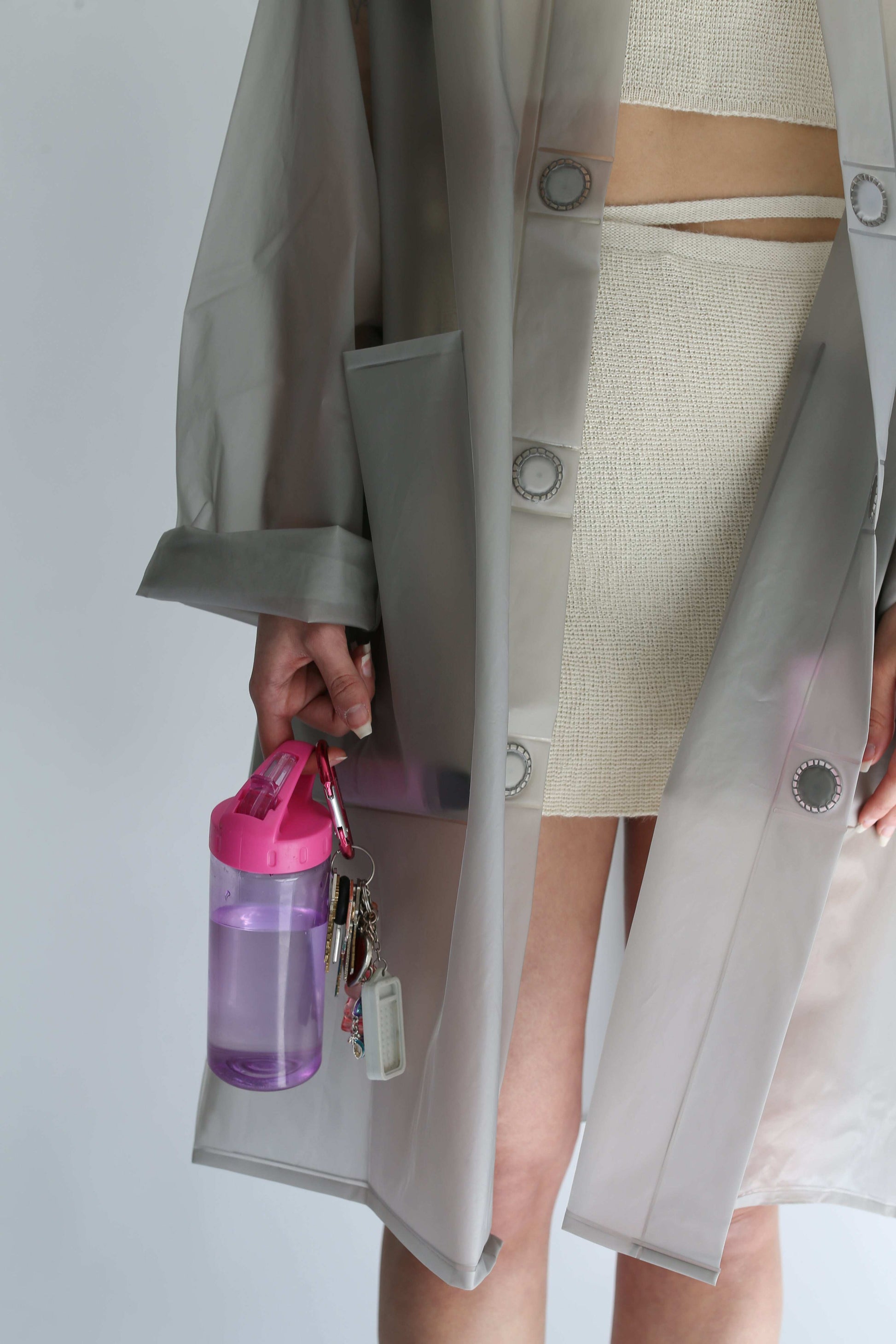 Detail of a woman wearing a transparent trench coat over a white knit skirt, holding keys and a bottle 