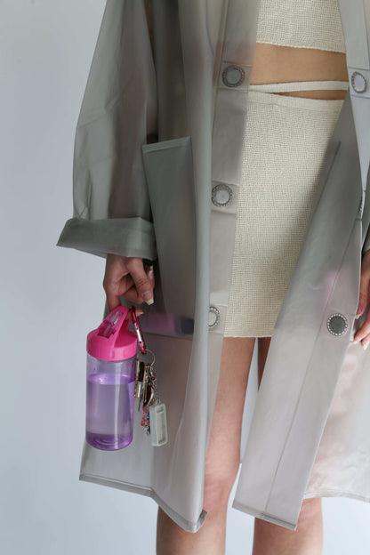 Detail of a woman wearing a transparent trench coat over a white knit skirt, holding keys and a bottle 