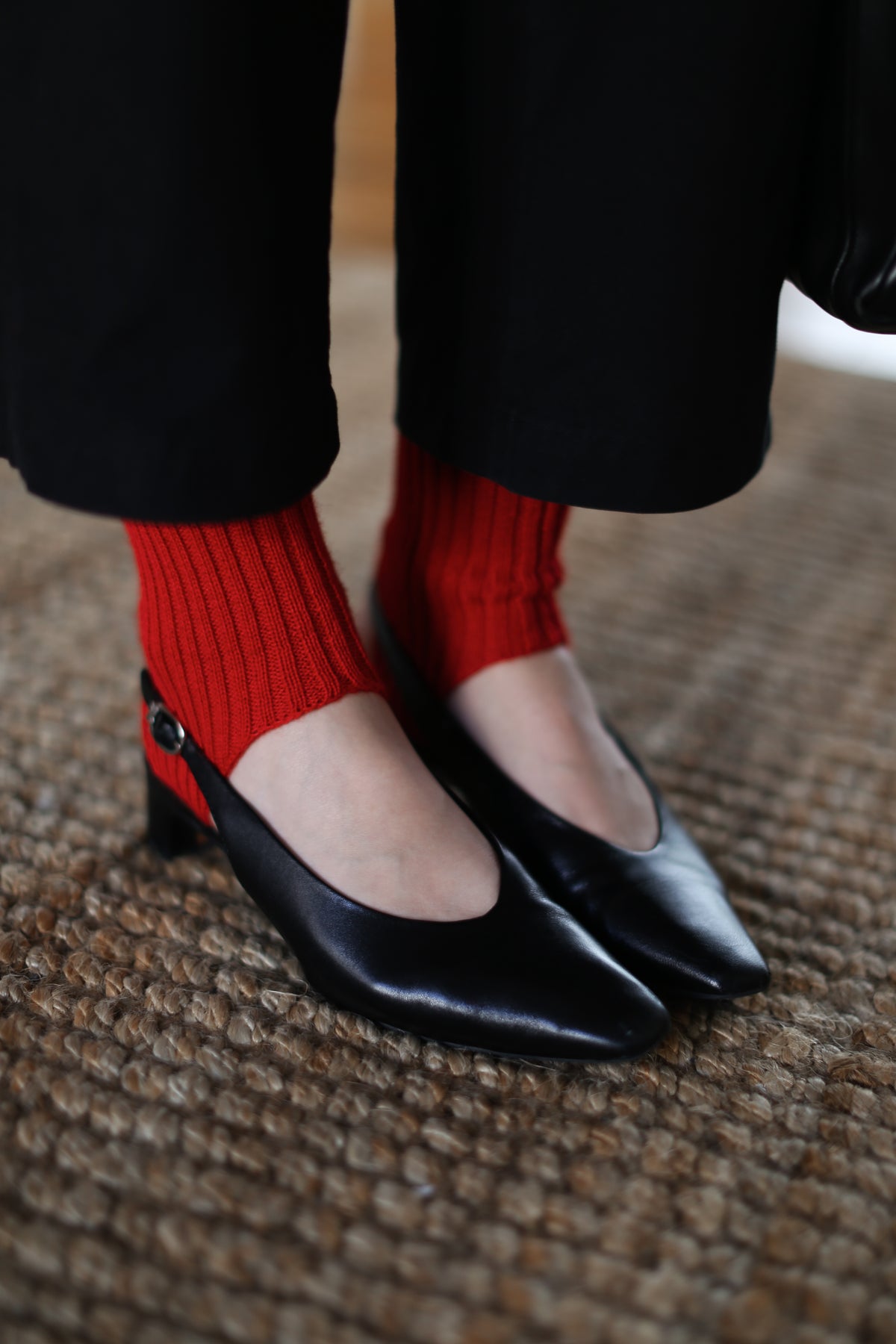 A close-up of a person's feet wearing black leatherflats and bright red ribbed leg warmers standing on a brown woven jute rug.