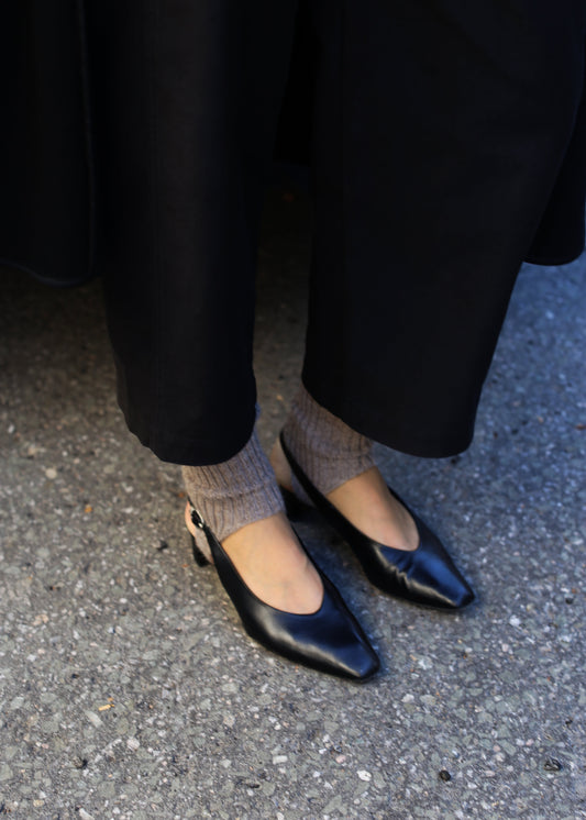 A close-up of a person's feet wearing black leather flats and taupe red ribbed leg warmers standing on a gray floor.