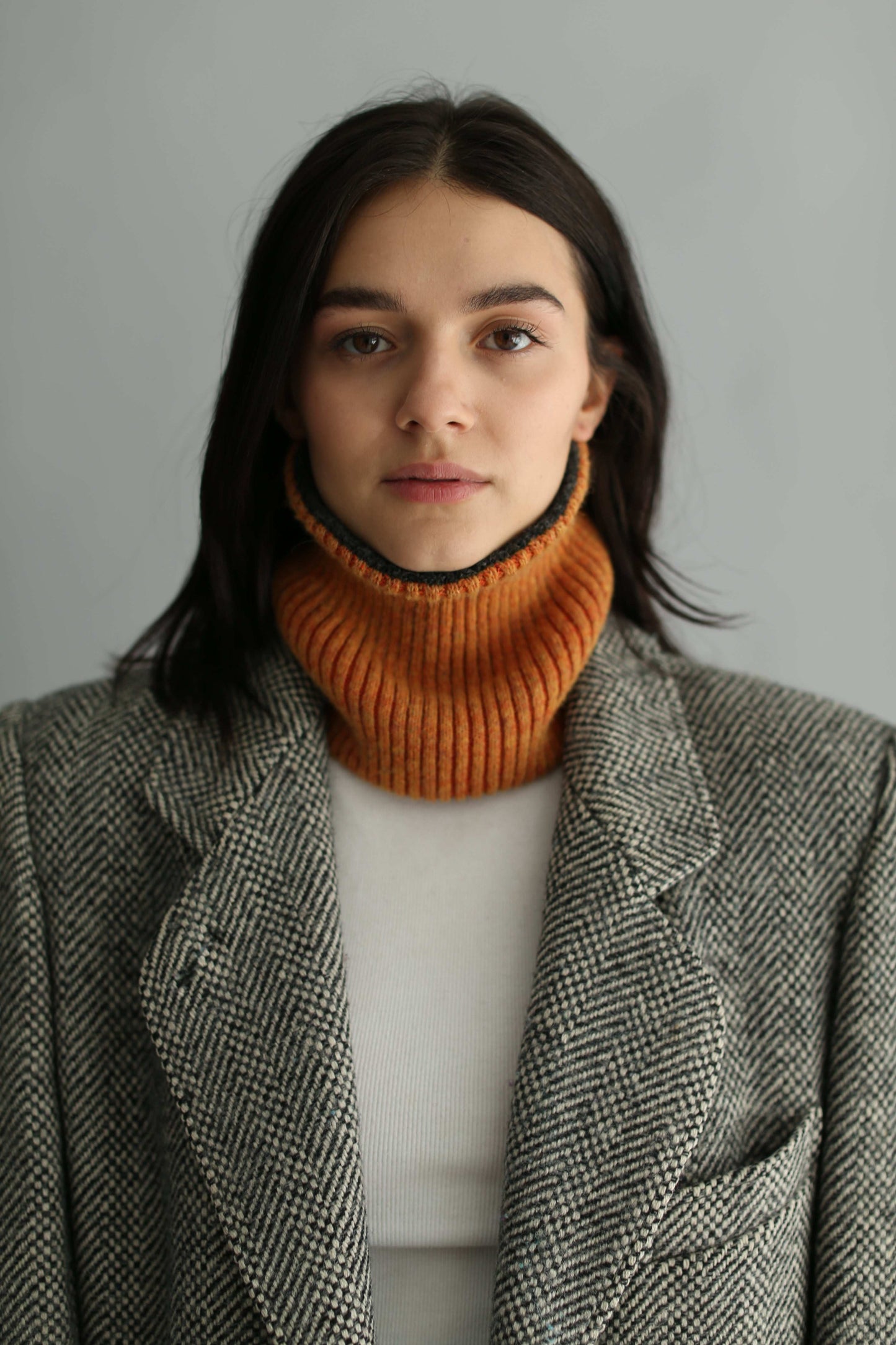 Portrait of a woman wearing an orange neck warmer, a tweed grey jacket and white sweater on a white background