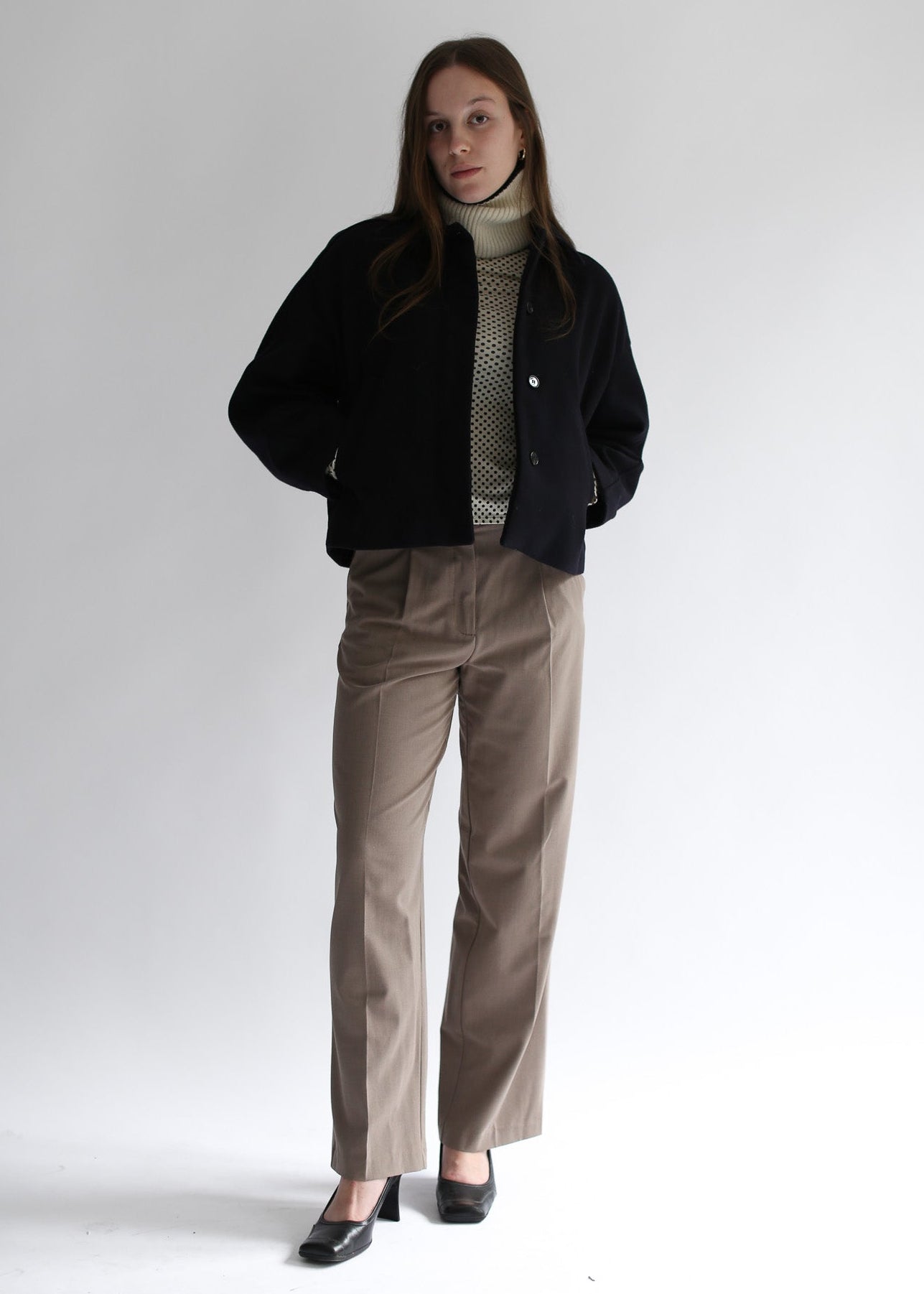 Model wearing a white neck warmer, a black jacket, beige sweater, and brown pants on a white background