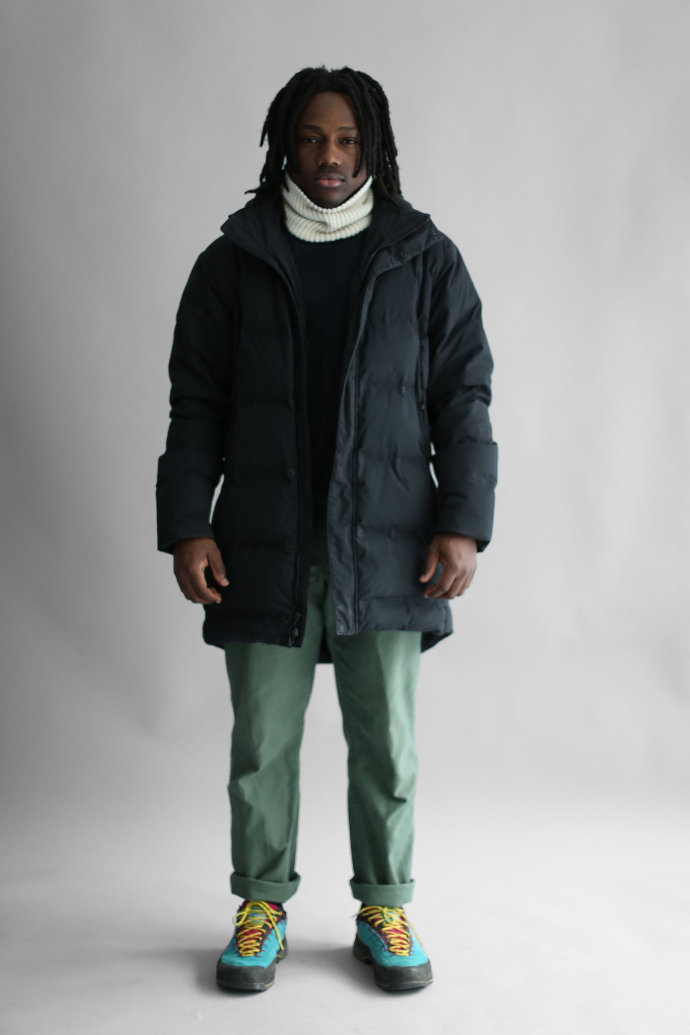 Model wearing a white neck warmer, a black puffa and green pants on a white background