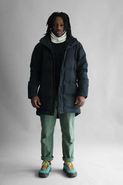 Model wearing a white neck warmer, a black puffa and green pants on a white background