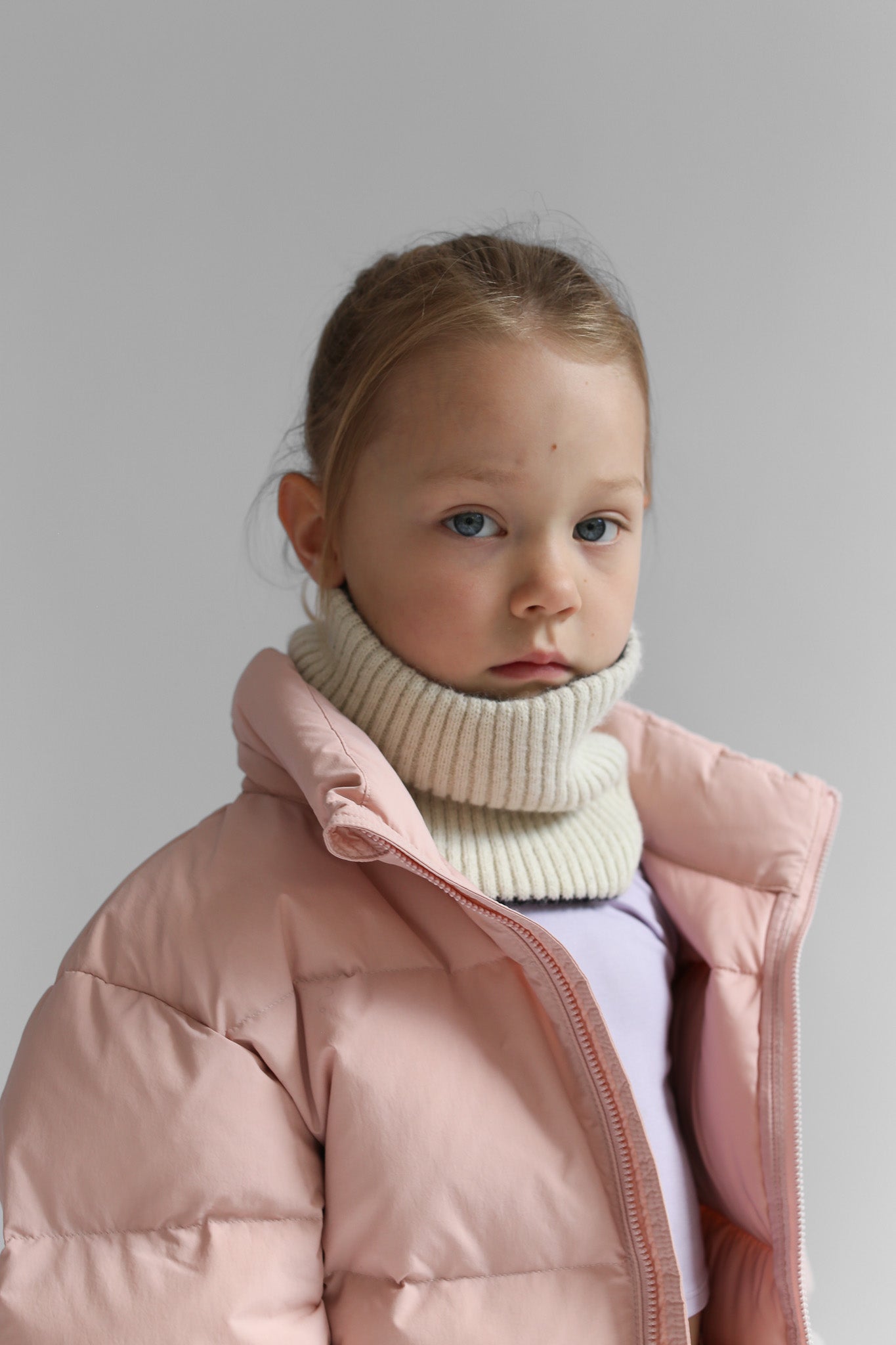 Child wearing a pink puffer jacket and white neck warmer against a plain background