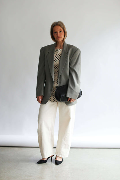 Model wearing a grey oversize jacket white pants and a white polka dot top and skirt on a neutral background