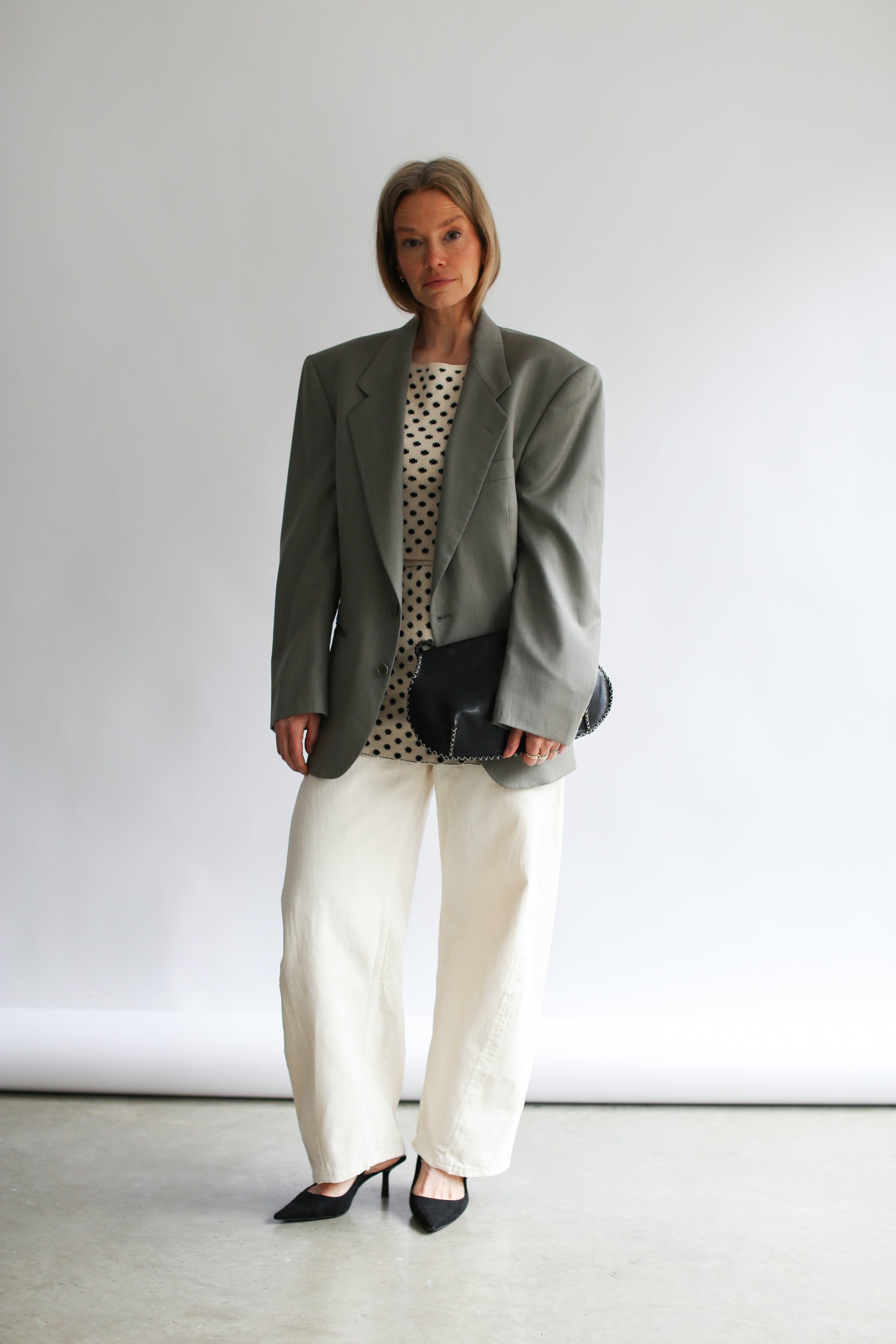 Model wearing a grey oversize jacket white pants and a white polka dot top and skirt on a neutral background