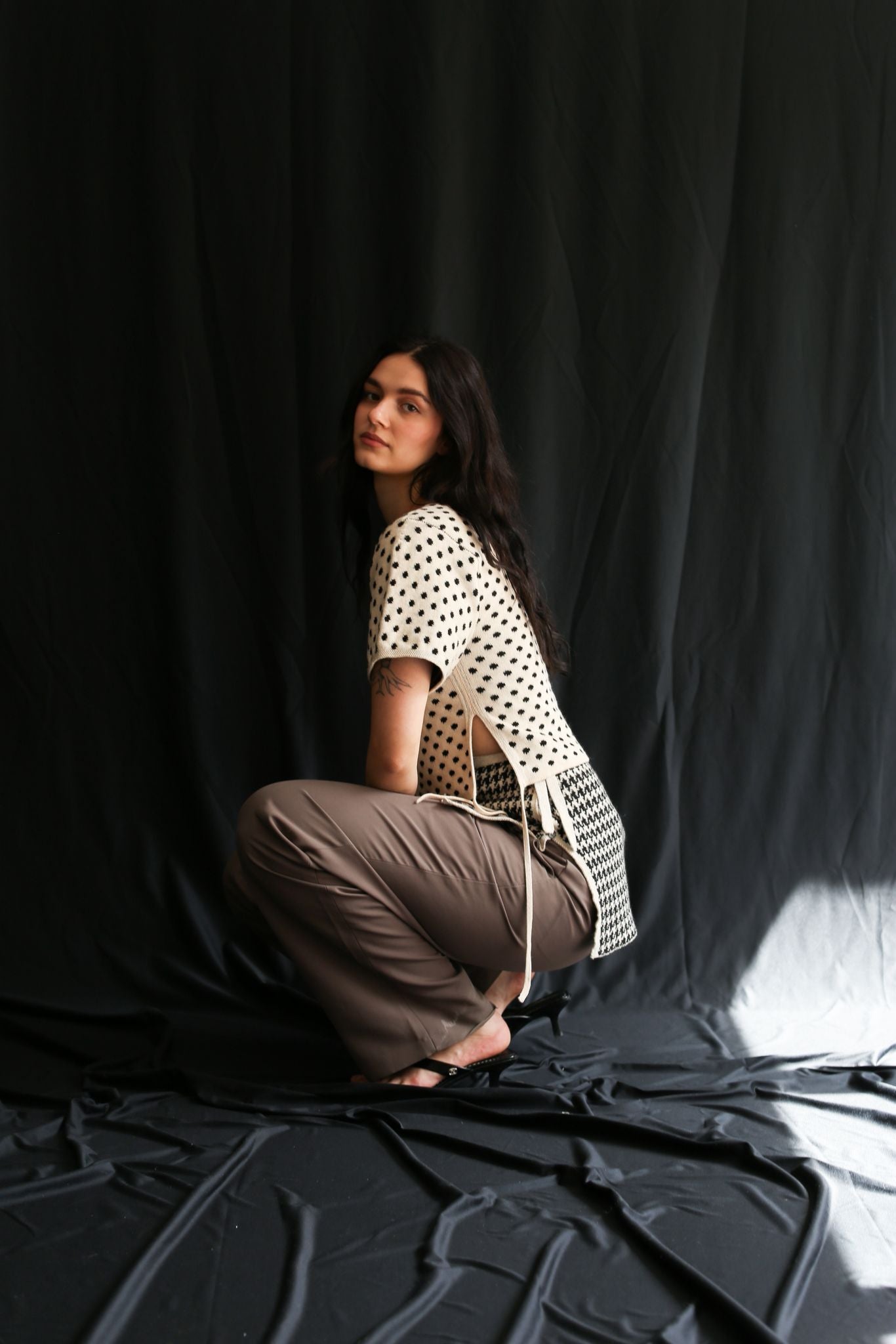 Woman sit, wearing a knitted polka dot top and a houndstooth skirt against a black background