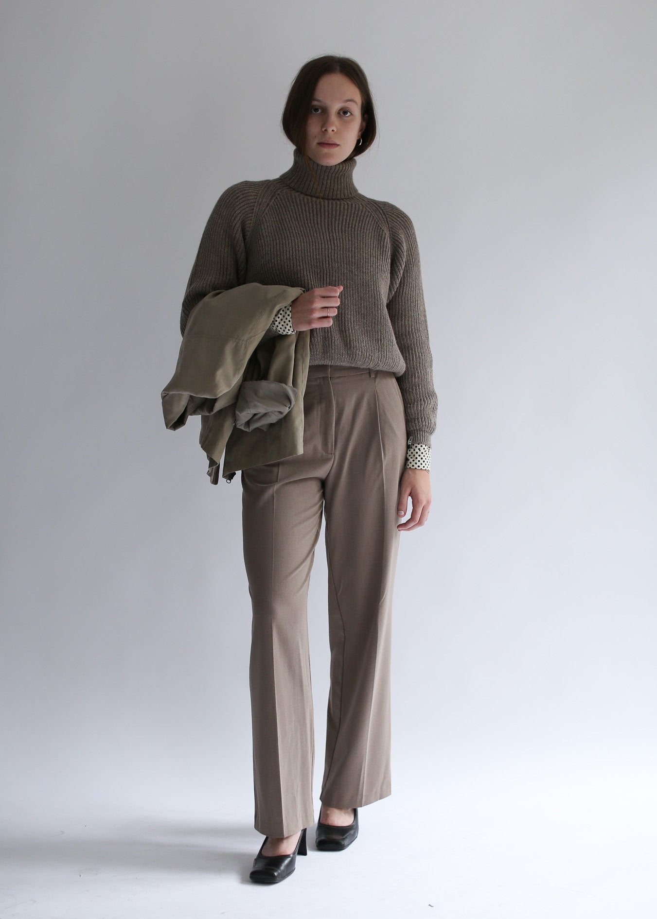 Woman wearing a brown cropped turtleneck sweater and beige pants, holding a jacket against a white background.
