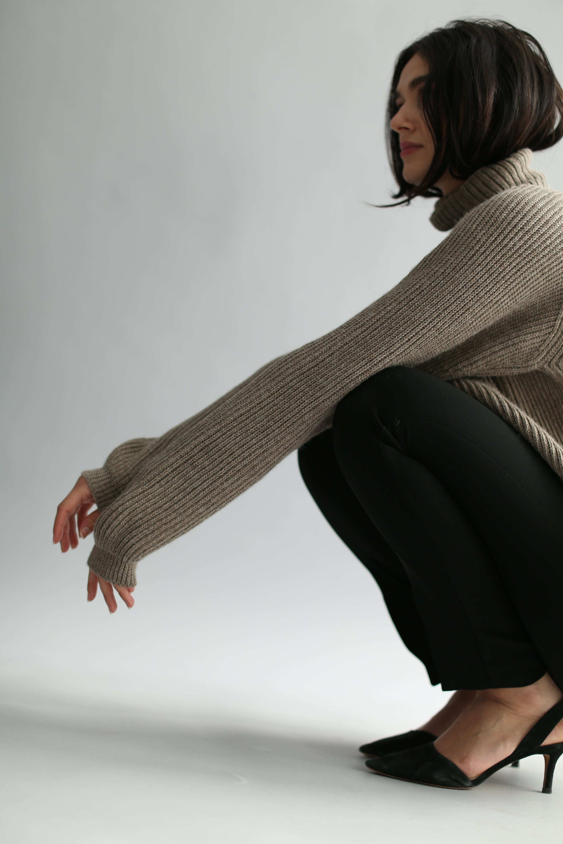 Woman squatting, wearing a brown ribbed turtleneck sweater and black pants against a gray background