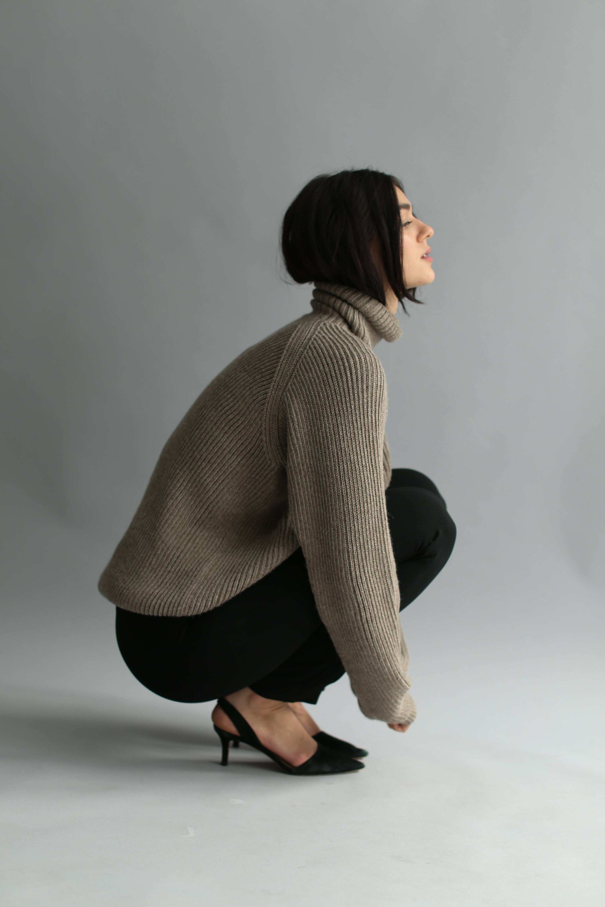 Woman squatting, wearing a brown ribbed turtleneck sweater and black pants against a gray background