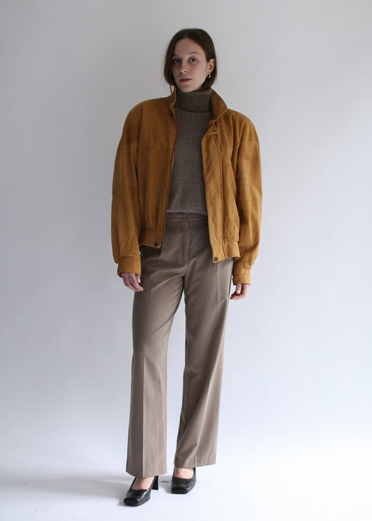 Model wearing a brown ribbed turtleneck, mustard suede jacket and beige pants against a white background