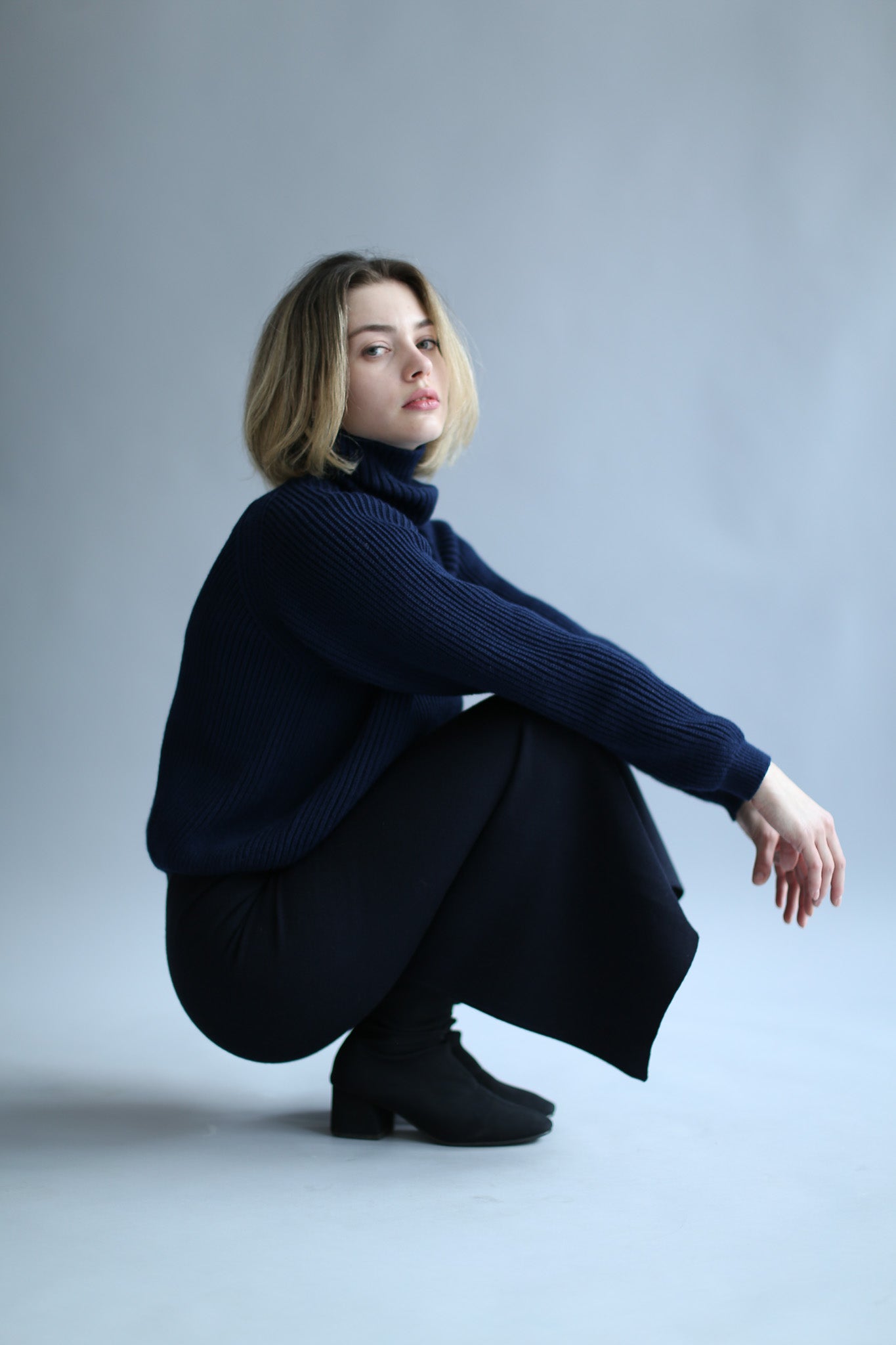 Model squatting wearing a navy ribbed turtleneck and black pants against a white background