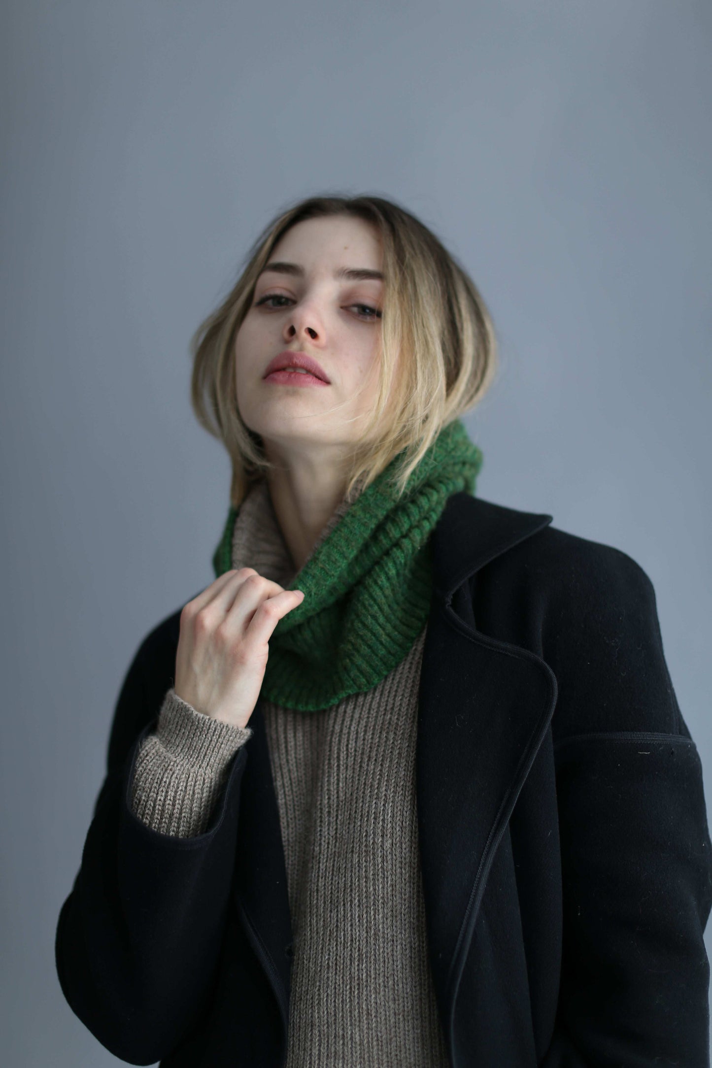 Portrait of a blonde woman wearing a green balaclava, black coat and brown sweater against a white background