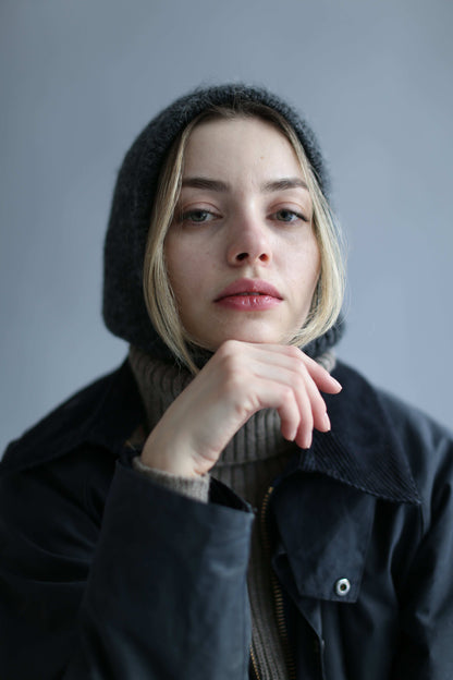Portrait of a blonde woman wearing a grey balaclava, black waxed jacket and brown sweater against a white background