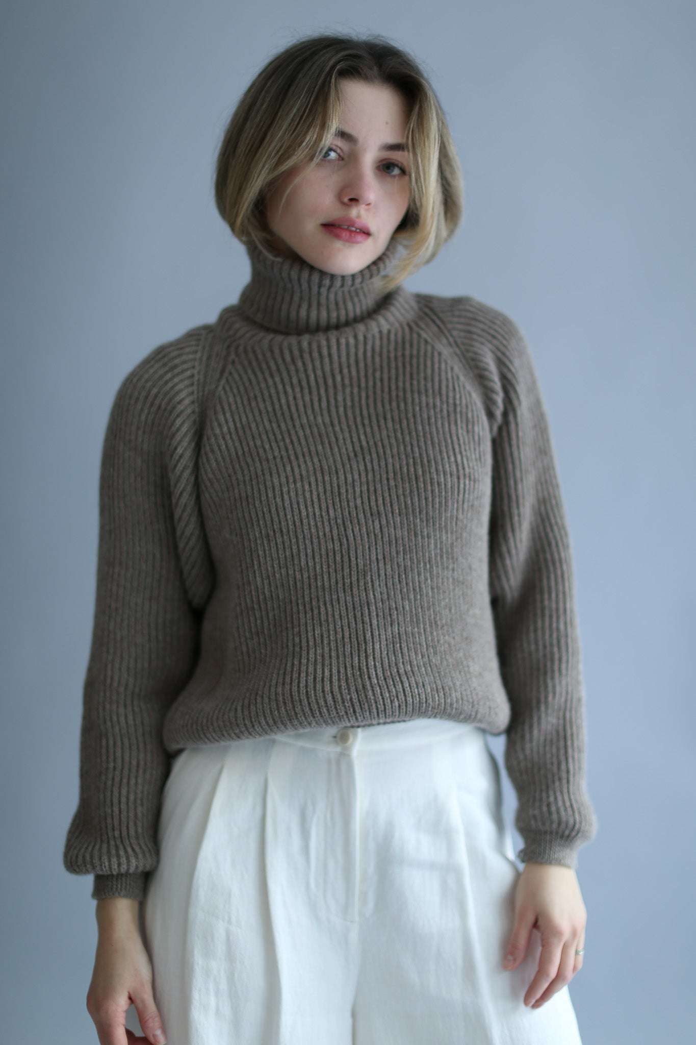 A model wearing a brown ribbed turtleneck and white pants against a white background