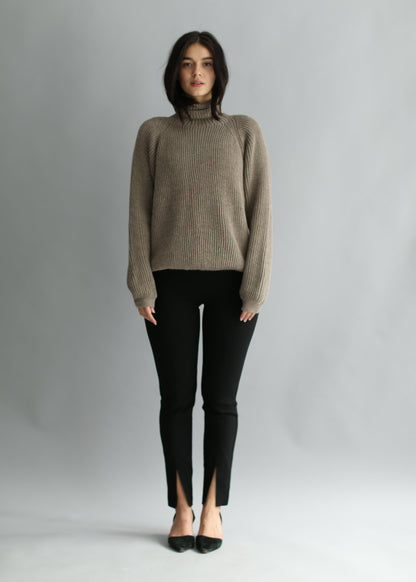 Woman wearing a brown ribbed turtleneck sweater and black pants against a gray background