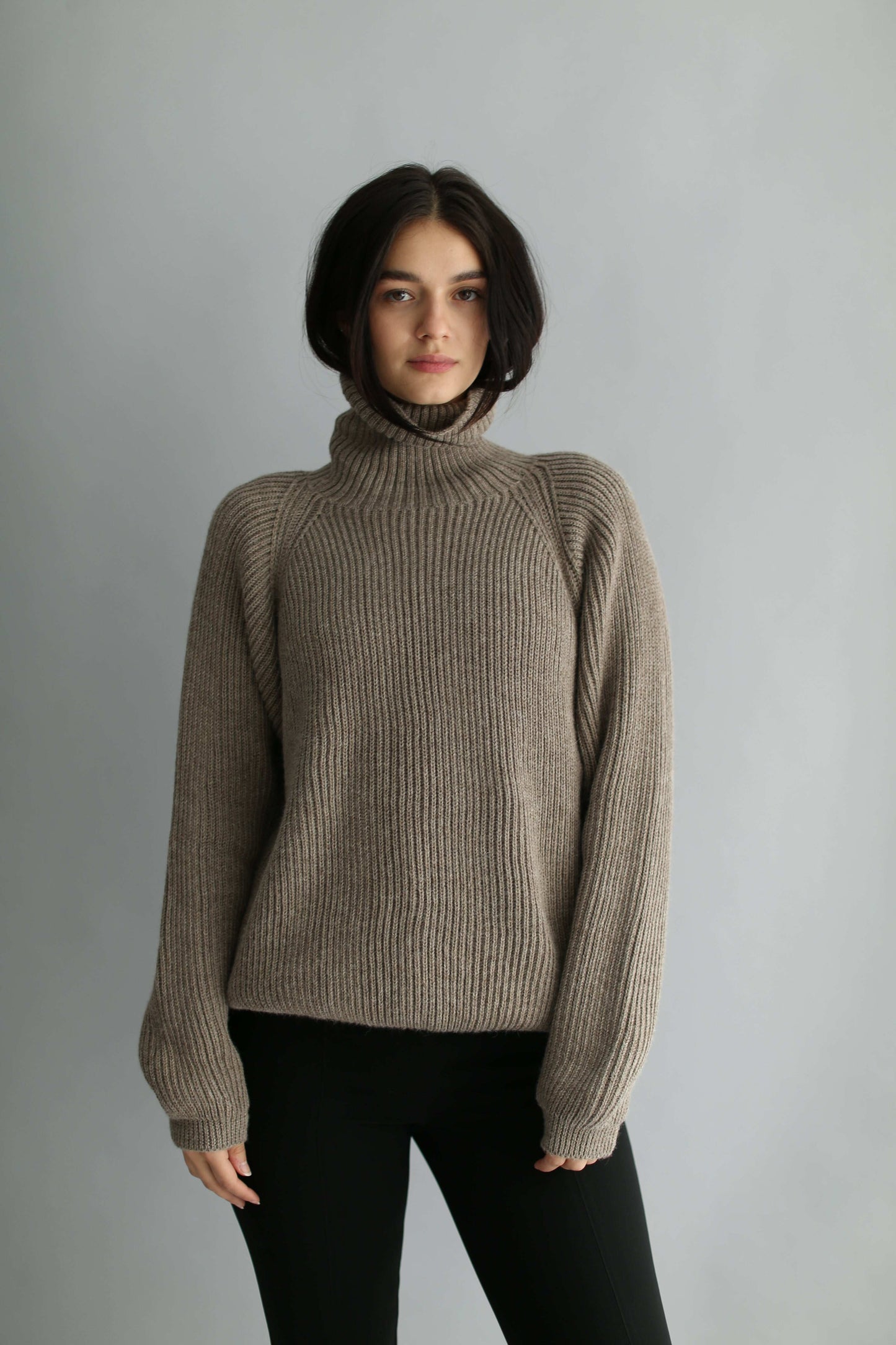 Woman wearing a brown ribbed turtleneck sweater and black pants against a gray background