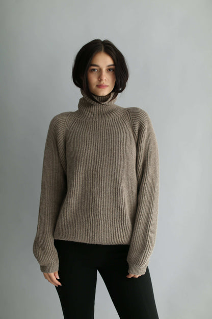 Woman wearing a brown ribbed turtleneck sweater and black pants against a gray background