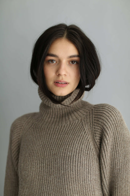 Portrait of a woman wearing a brown ribbed turtleneck sweater and black pants against a gray background